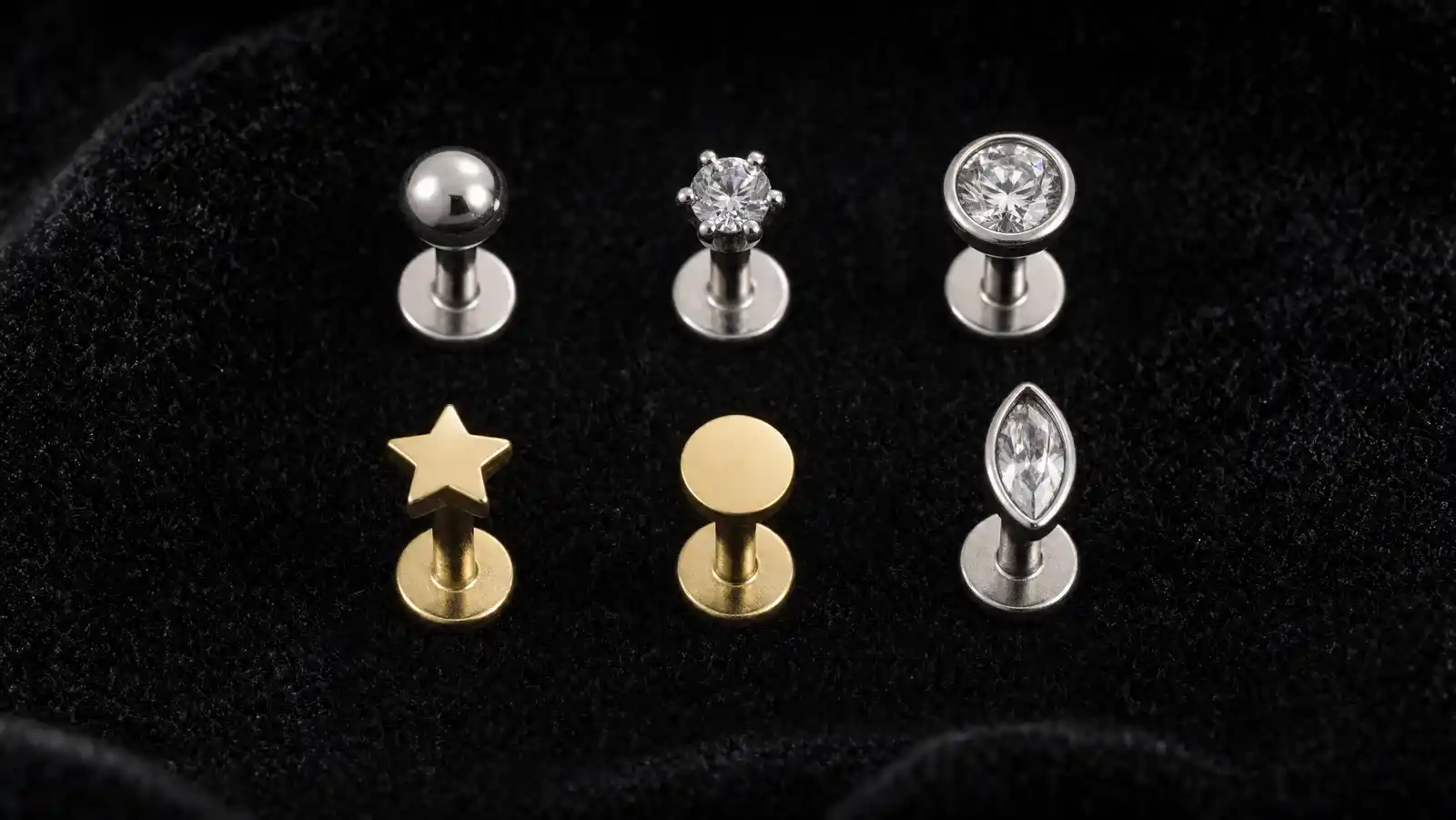 Six titanium flat back stud earrings standing upright on their flat discs in two rows on black velvet: a polished dome, a prong-set round stone, a bezel-set round stone, a gold star, a plain gold disc and a marquise-cut stone