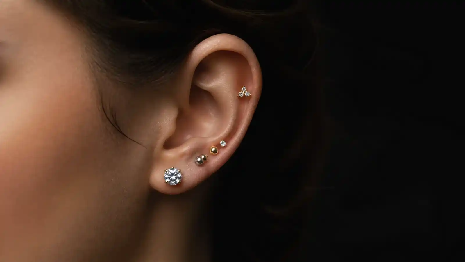 A curated ear in three-quarter profile against a black background: a larger round solitaire stud in the lobe, two smaller graduated studs in mixed gold and silver above it, and a gold three-stone cluster higher on the upper ear