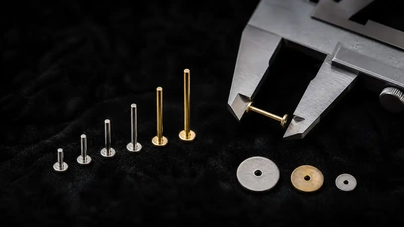 Six titanium and gold flat back earring posts arranged in ascending length on black velvet beside steel calipers measuring a gold post, with three flat disc backs of different diameters in the foreground