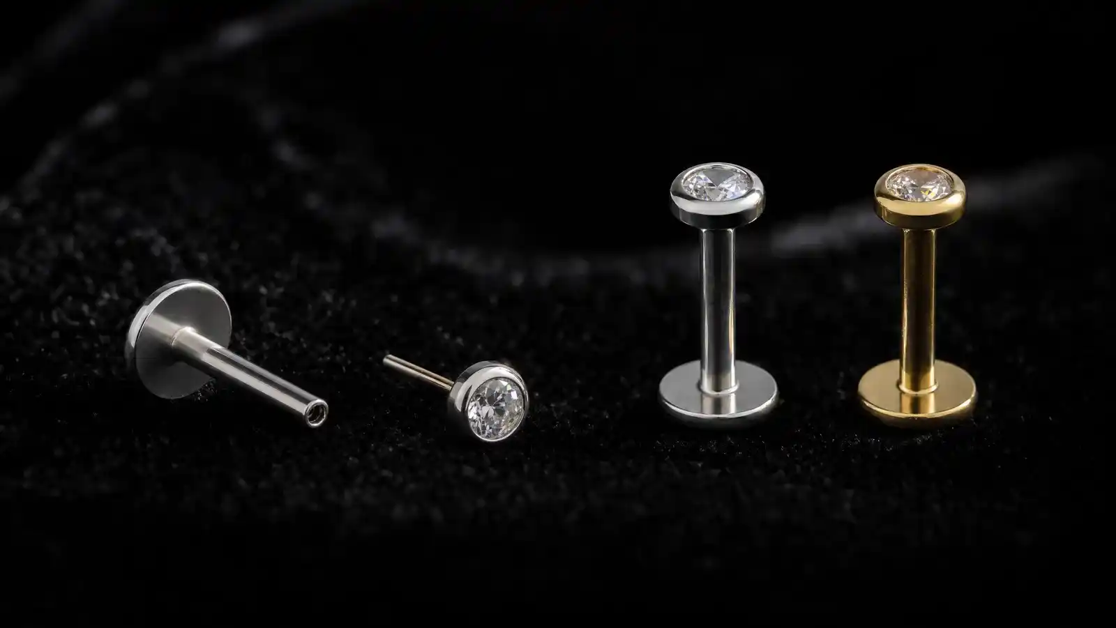 Four flat back labret earrings on black velvet: a bare titanium flat disc back with its hollow post lying on its side, a loose push-pin top set with a round clear stone, and two assembled studs standing upright on their discs, one silver titanium and one gold