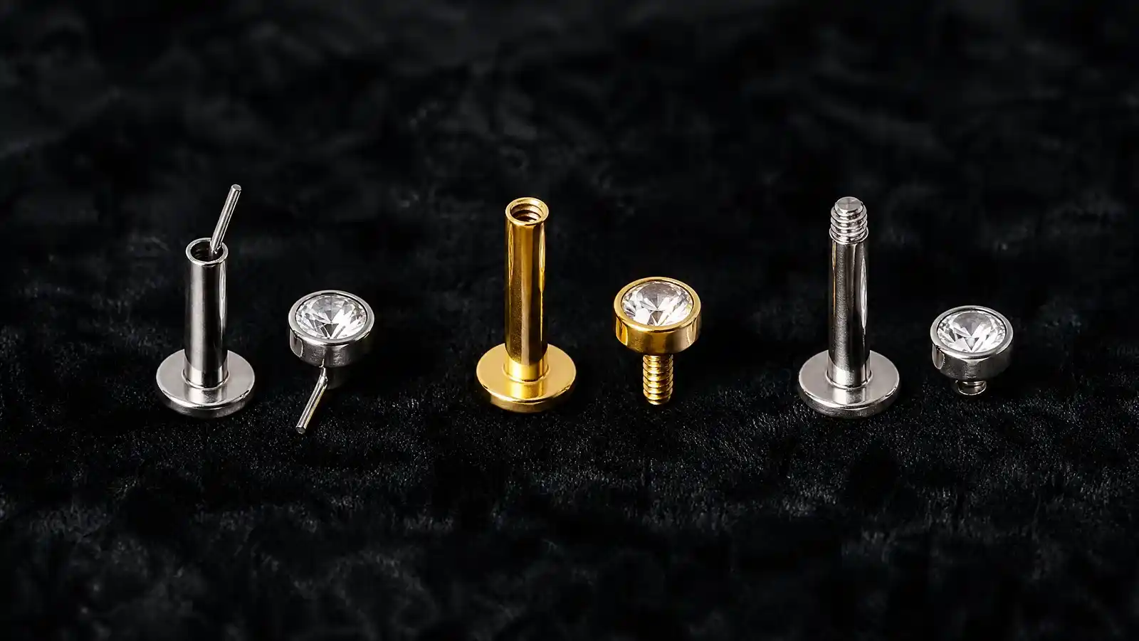 Three flat back earring systems disassembled in a row on black velvet: a silver threadless post with its bent-pin top, a gold post with a tapped internal bore beside a top carrying a male screw thread, and a silver post with threads cut on the outside of the bar